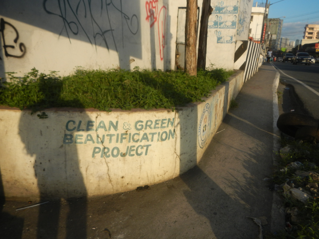 Eine Straße mit Fahrzeugen, Gebäuden, Bäumen, Polen, Drähten und einer Wand mit Graffiti "Saubere und grüne Verschönerung" mit Pflanzen und Gras im Vordergrund und einem bewölkten Himmel im Hintergrund.