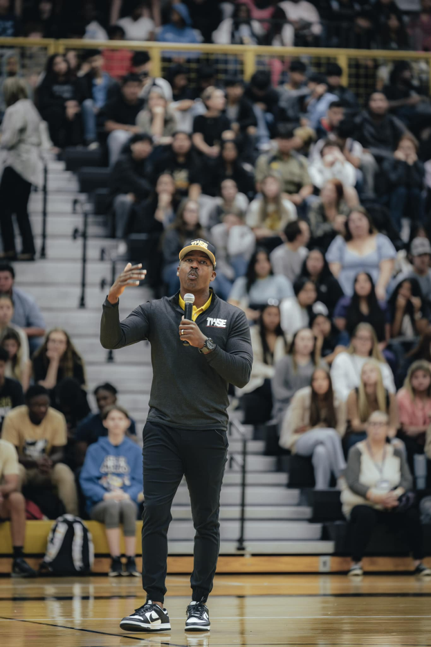 Ein Mann mit Mütze hält ein Mikrofon auf einem Basketballfeld, mit einer Gruppe von Menschen auf den Stufen und Taschen auf dem Boden hinter ihm.