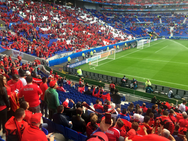 Eine große Menschenmenge in einem Stadion schaut bei einem Fußballspiel zu, mit Menschen auf Stühlen und Stehplätzen, Bannern, einem Tor und Deckenleuchten.