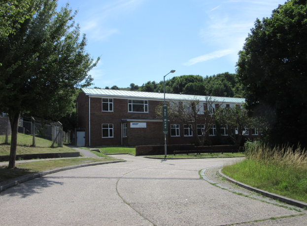Ein Schulgebäude mit Fenstern, einem Straßenpfahl, einem Schild, einer Baumgruppe, Gras, einem Zaun und einer bewölkten Himmel, das in der Mitte eines Parks umgeben von saftigem Gras liegt.
