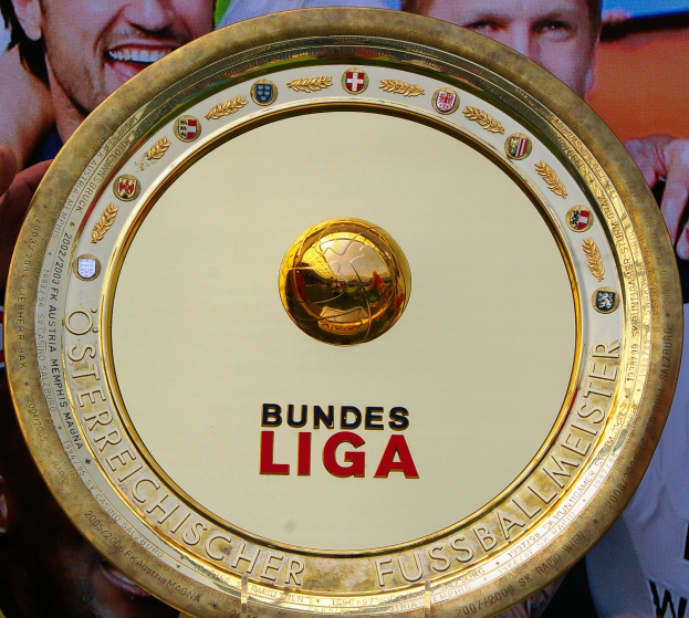 Mann hält goldenen Schild mit 'Bundesliga'-Text hoch, im Hintergrund ein Banner mit drei Personen