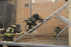 Feuerwehrleute in Helmen und Luftzylindern klettern an einer Leiter vor einem Backsteingebäude mit Rohren auf dem Boden und einer Metallstange unten, mit einem anderen Gebäude mit Fenstern und Netz im Hintergrund.
