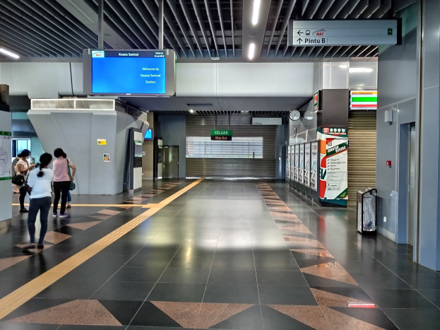 Menschen gehen durch einen Bahnhof mit einem Wagen auf der rechten Seite, Deckenleuchten, einer Uhr an der Wand und Informationsanzeigen.