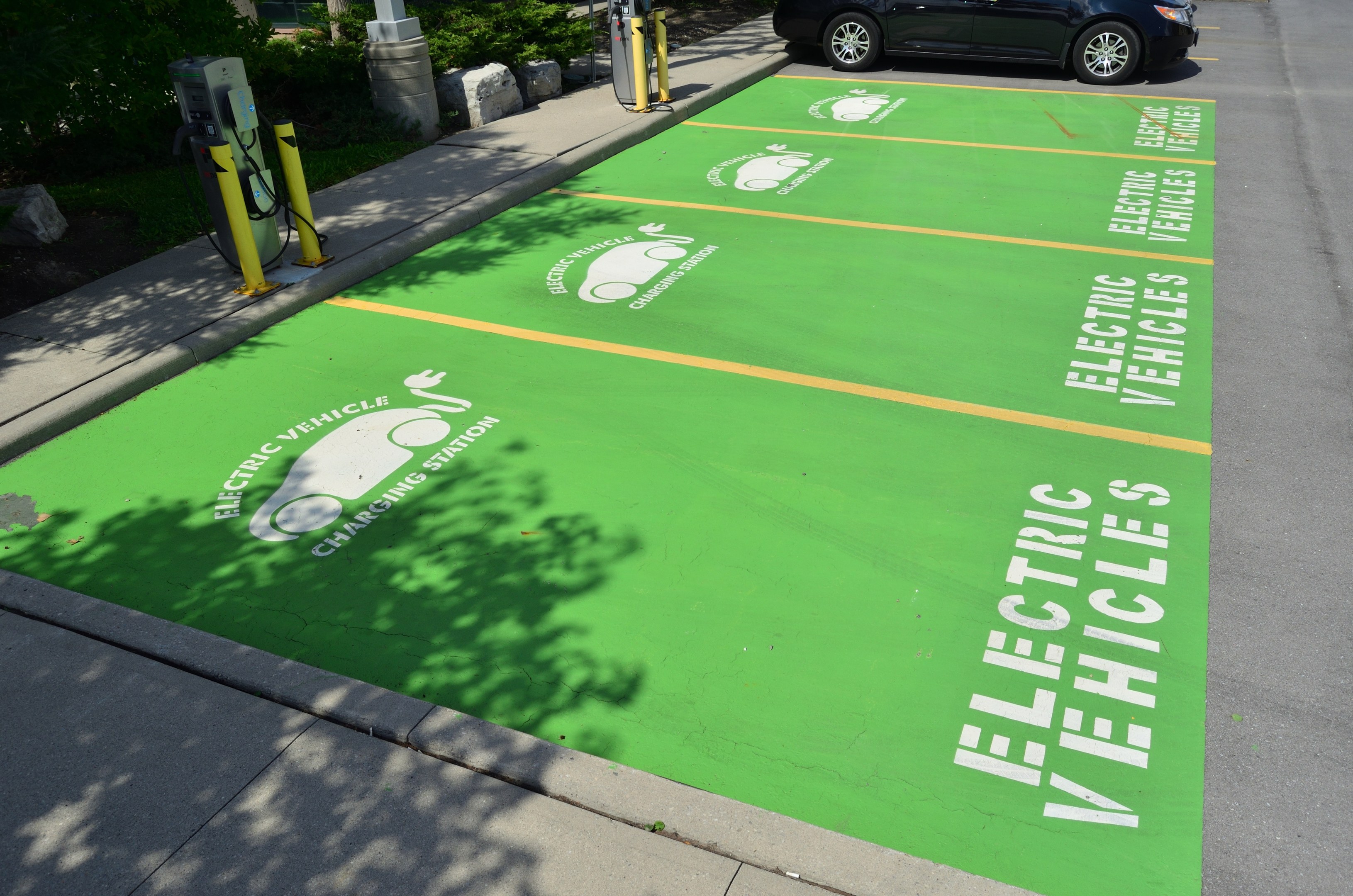 Elektroauto an einer Ladesäule mit grünem Bodenmatten markiert, umgeben von Bäumen, Pflanzen, Polen und anderen Gegenständen auf einem Parkplatz.