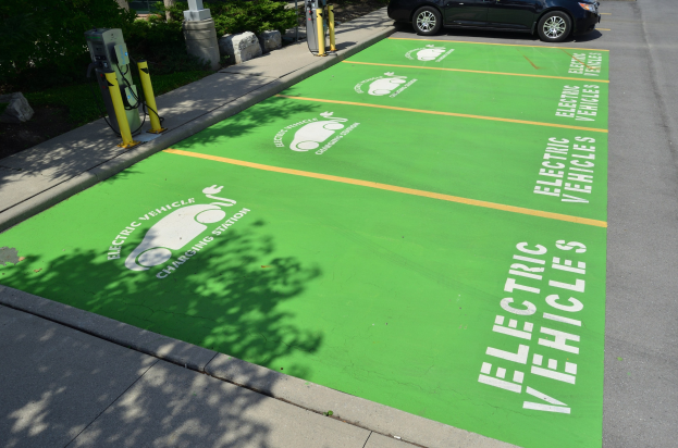 Elektroauto an einer Ladesäule mit grünem Bodenmatten markiert, umgeben von Bäumen, Pflanzen, Polen und anderen Gegenständen auf einem Parkplatz.