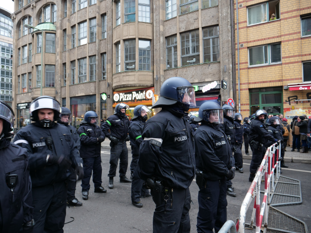 Eine Gruppe von Polizisten vor einer Menschenmenge hinter Absperrungen auf einer Straße mit Gebäuden, Schildern, Laternenmasten und einem klaren Himmel im Hintergrund.