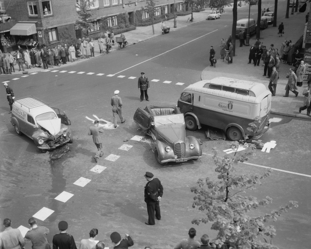 Schwarz-weiß-Szene eines Autounfalls an einer Straßenkreuzung mit mehreren Fahrzeugen, darunter ein Lieferwagen, und Menschen, die sich darum versammeln; Gebäude, Bäume, Pfosten und Texttafeln sind im Hintergrund sichtbar.