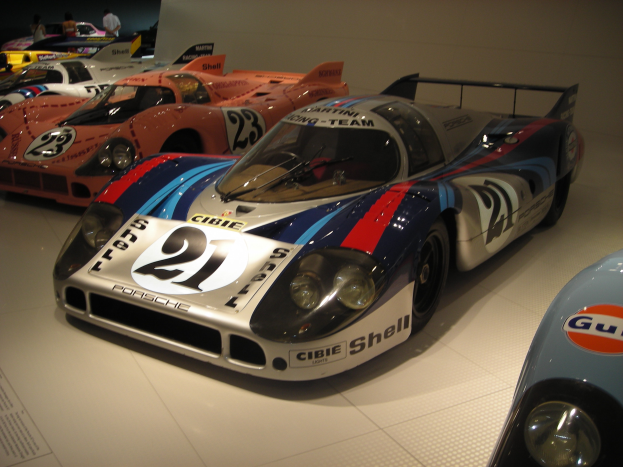 Ein Porsche 917K Le Mans-Rennwagen wird in einem Museum ausgestellt, umgeben von anderen Sportwagen und Menschen, mit einer Wand im Hintergrund und einem Blatt Papier auf der linken Seite.