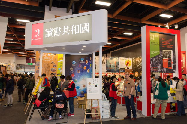 Eine große Gruppe von Menschen an einem Messestand mit Bannern, Texttafeln mit der Aufschrift "Book Republic Hong Kong Book Fair" und Deckenbeleuchtung.