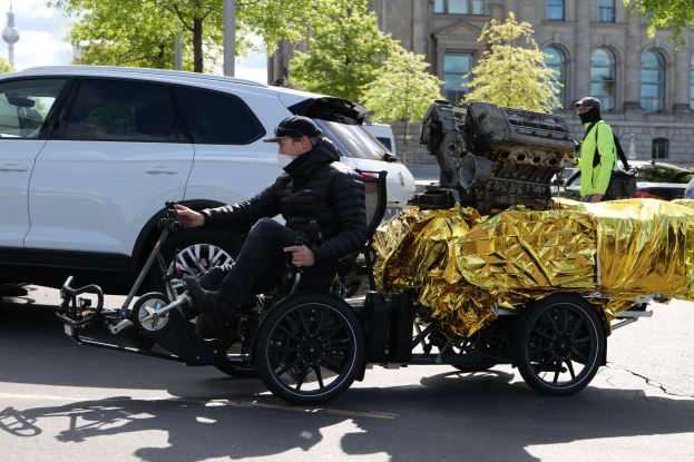 Ein Mann im Rollstuhl mit einem großen Motor auf dem Rðcken, umgeben von Fahrzeugen auf einer Straße mit Bäumen, Gebäuden und einem klaren blauen Himmel im Hintergrund, tragend eine schwarze Jacke, eine Kappe und haltend ein Objekt.