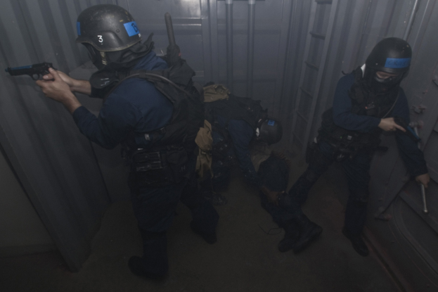 Eine Gruppe von Polizisten in Einsatzausrüstung, einer mit einer Waffe in der Hand, neben einem Metallcontainer und einer Leiter in einem beengten Raum.