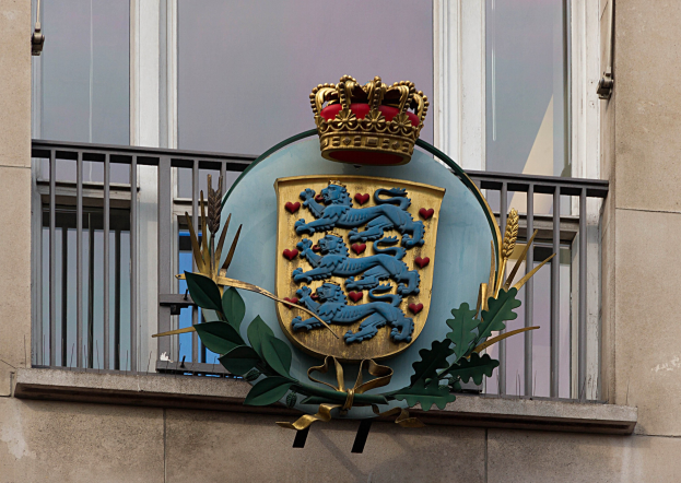 Wappen der britischen Königsfamilie auf einem Gebäude gezeigt, begleitet von einem Logo mit Krone und Blättern, mit Fenstern und Gittern im Hintergrund.