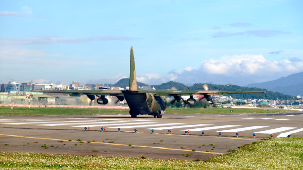 Ein großes Militärluftfahrzeug hebt von einer Flugplatzrollbahn mit Gras und kleinen Pflanzen ab, vor einem Hintergrund aus Gebäuden, Bäumen, Bergen und einem klaren blauen Himmel.