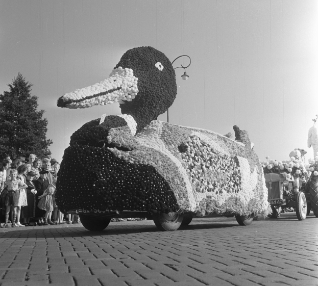 Ein Schwarz-Weiß-Foto eines mit Blumen geschmückten Entenwagens, umgeben von Menschen auf der linken Seite, mit Bäumen und einem klaren Himmel im Hintergrund.