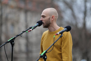 Ein kahlkopfiger Mann in einem gelben T-Shirt, der in ein Mikrofon singt, vor einem unscharfen Hintergrund.