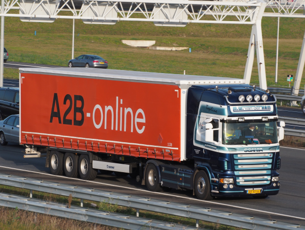 Ein roter und blauer Sattelschlepper, der auf einer Autobahn neben einer Brücke mit Geländer fährt, umgeben von grünem Gras, Pfählen und Gebäuden im Hintergrund.
