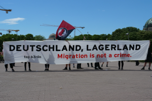 Eine Gruppe von Menschen, die eine Fahne hält, auf der 'Deutschland, Lagerland Migration ist kein Verbrechen' steht, und eine Flagge, mit Straßenlaternen, Bäumen, Kränen, einem Gebäude und einem bewölkten Himmel im Hintergrund.