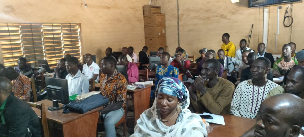 Eine Gruppe von Menschen in einem Klassenzimmer mit Monitoren, Taschen, Papieren und Büchern auf den Tischen, einem Fenster auf der linken Seite und Kartons und einem Fernseher an der Wand, die an einer Techniktrainingssitzung teilnehmen.
