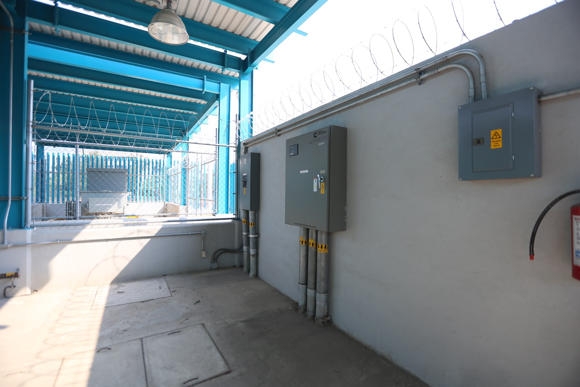 Ein Raum mit einem Feuerlöscher auf der rechten Seite, elektrischen Schaltkästen und Rohren an der Wand, einer Deckenlampe und einem Zaun im Hintergrund, was auf eine elektrische Umspannstation hinweist.