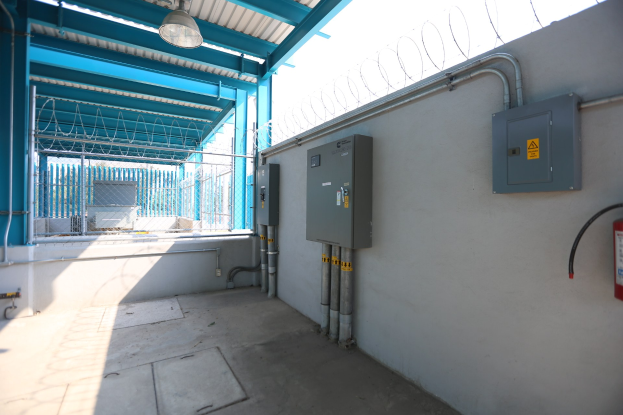 Ein Raum mit einem Feuerlöscher auf der rechten Seite, elektrischen Schaltkästen und Rohren an der Wand, einer Deckenlampe und einem Zaun im Hintergrund, was auf eine elektrische Umspannstation hinweist.