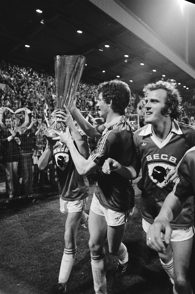 Eine Gruppe von Männern, die einen Pokal auf einem Fußballfeld halten, umgeben von einer Menge hinter einem Zaun, die ihren Sieg im FA Cup Finale feiert, dargestellt in Schwarz-Weiß.