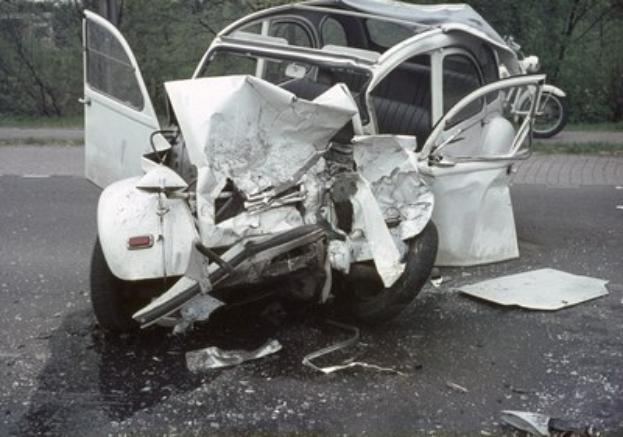 Ein schwerbeschädigtes Auto mit eingedellter Frontpartie am Straßenrand, umgeben von Bäumen und einem entfernten Gebäude, mit einem weiteren Fahrzeug im Hintergrund.