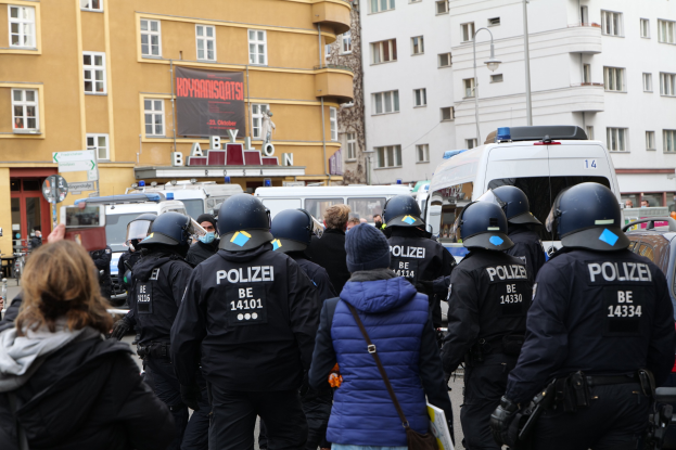 Polizeibeamte in Uniform vor einer Menge bei einer Demonstration in Berlin, mit Fahrzeugen, Gebäuden und einem Banner im Hintergrund.