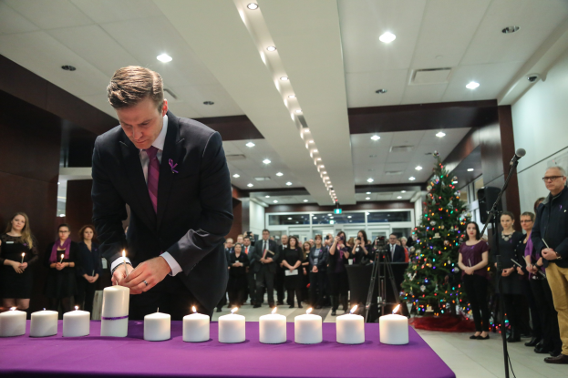 Ein Mann im Anzug zündet Kerzen auf einem Tisch an, während Menschen drumherum stehen, mit einem Mikrofon, einem Weihnachtsbaum und Deckenleuchten im Hintergrund.