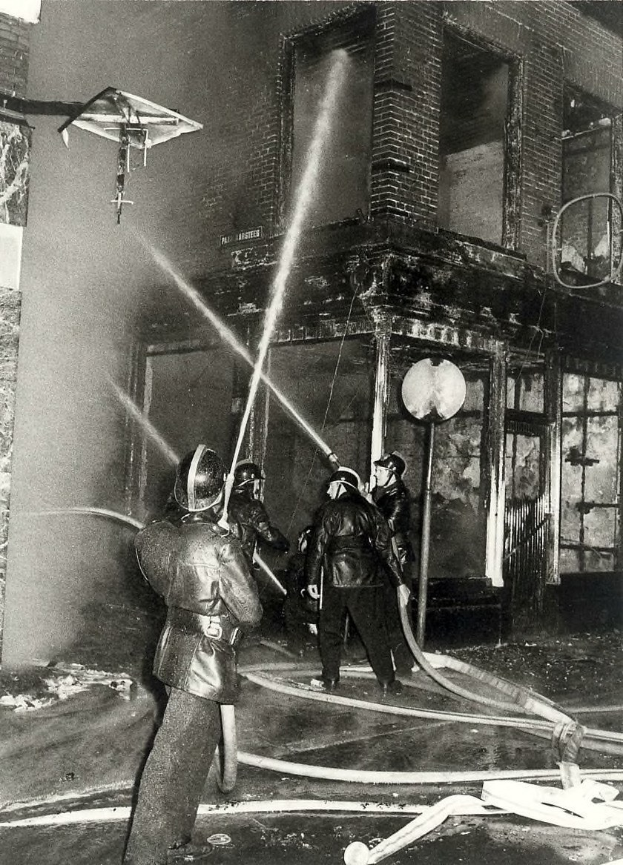 Schwarz-weiß-Foto von Feuerwehrmännern in Helmen mit Schläuchen in der Hand, die vor einem feuerbeschädigten Gebäude stehen.