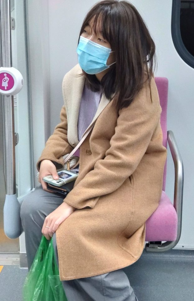 Eine Frau mit Gesichtsmaske sitzt in einem Zug, hält ein Mobiltelefon und eine Tasche und hat eine Tür links von ihr.