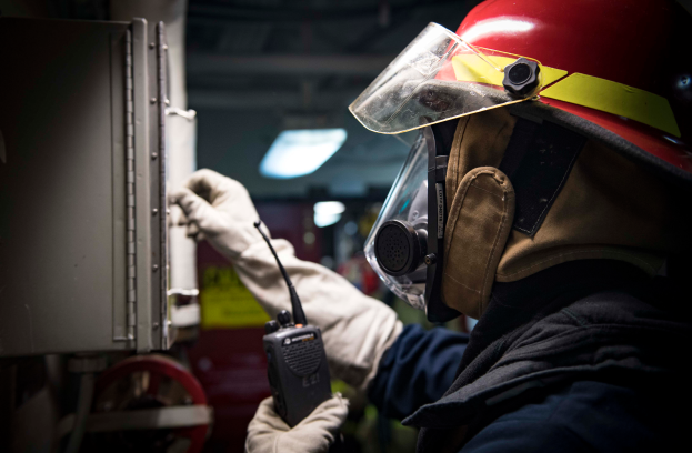 Ein Feuerwehrmann in voller Ausrüstung hält ein Funkgerät und ein metallisches Objekt, mit einem unscharfen Hintergrund.