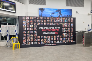 Eine große Wand mit mehreren Bildern, einem gelben Stuhl, israelischen Regierungsflaggen auf Stangen, einer Tafel mit dem Text "Bring Them Home Now", einem montierten Bildschirm, Deckenleuchten und anderen Gegenständen.