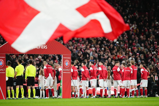 Eine Gruppe von Menschen auf einem Fußballfeld mit einer roten und weißen Flagge im Vordergrund, einem Bogen mit der Aufschrift "Bayern Munich vs. Bayern Munich Wetten & Vorschau" im Hintergrund und einer großen Menge im Stadion.