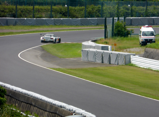Rennwagen auf einer Rennstrecke mit einem Polizeiwagen im Hintergrund, umgeben von Gras, Pflanzen, Bäumen und Absperrungen, mit einem Fahrzeug auf der rechten Seite und Text unten.