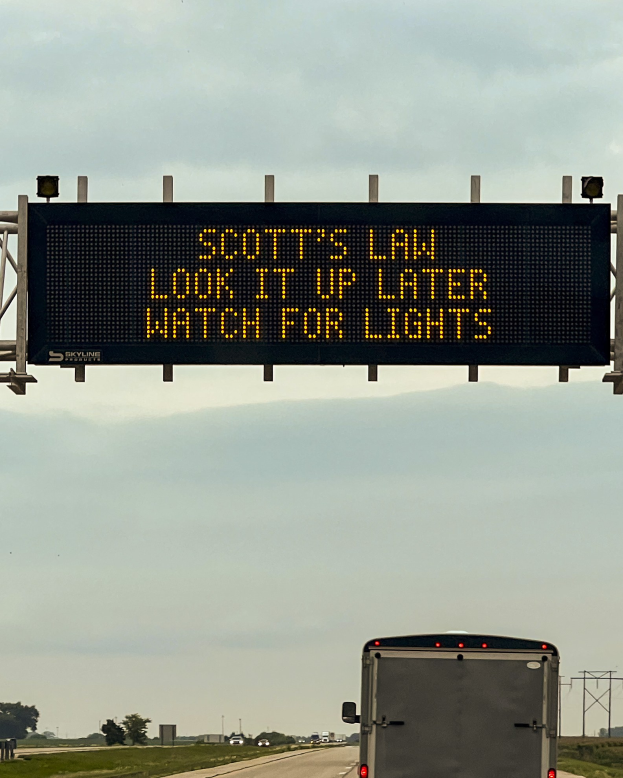 Ein Lkw fährt auf einer Autobahn mit einem Schild an der Seite, auf dem "Scott's Law: Schau es später nach, achte auf Lichter" steht, umgeben von Gras, Bäumen und Pfählen, mit dem Himmel im Hintergrund.