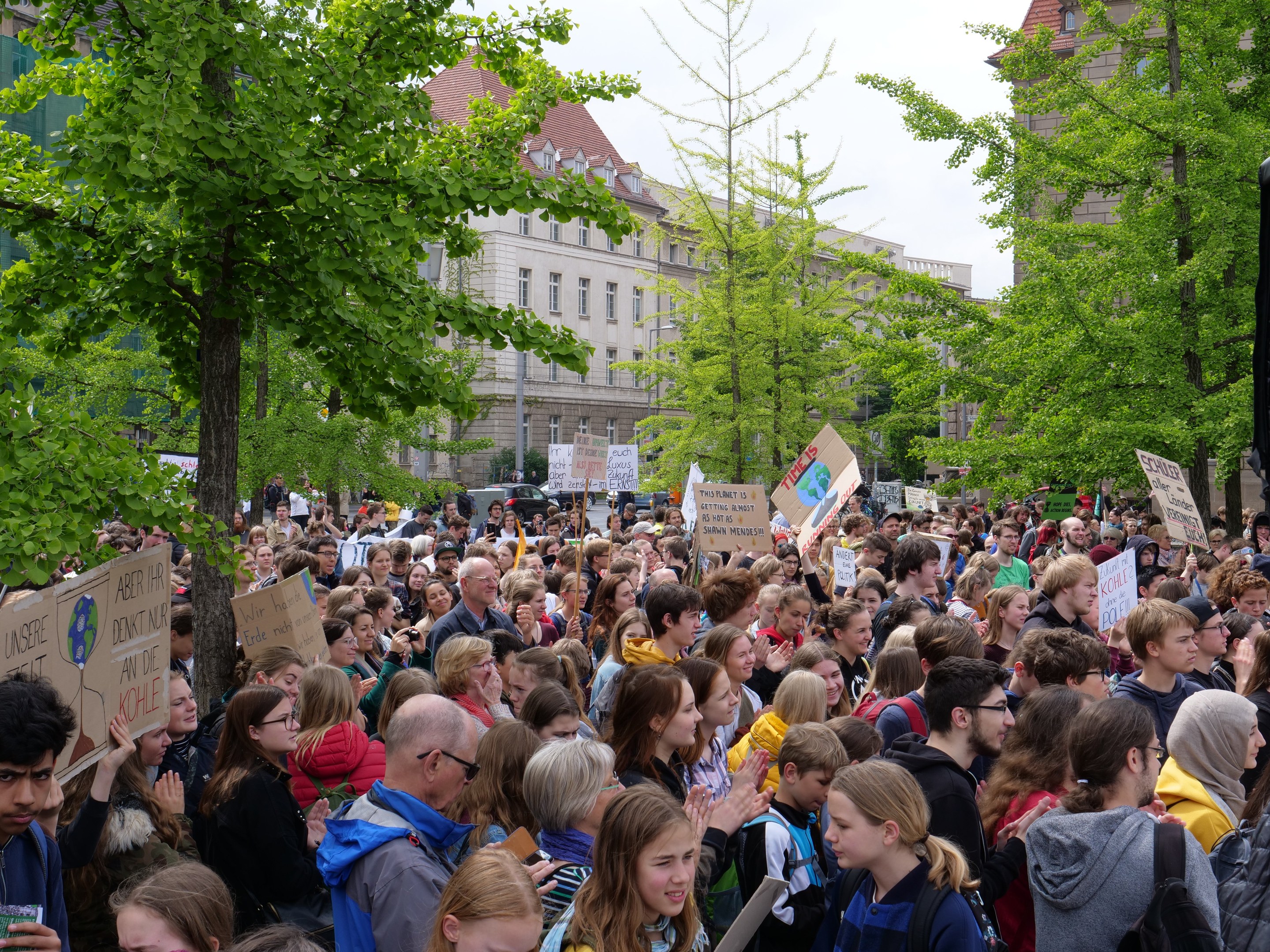 Eine große Menschenmenge protestiert vor einem Gebäude in Berlin und hält Schilder hoch, mit Bäumen, Fahrzeugen, einem Lautsprecher und dem Himmel im Hintergrund.
