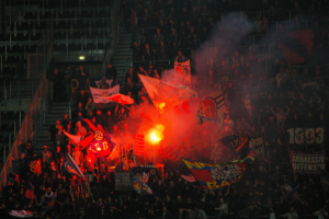 Eine große Menschenmenge in einem Stadion mit Fahnen und Bannern, beleuchtet von Scheinwerfern, umgeben von Stufen und Geländern, mit einem Feuer in der Mitte.