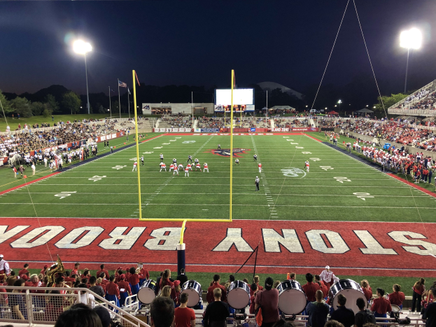 Ein nächtliches Fußballspiel in einem Stadion mit Spielern auf dem Feld, sitzenden Zuschauern und Musikern; ein Metallzaun umgibt das Gebiet, mit Laternen, Fahnen, einem Anzeigebildschirm, Bäumen, Hügeln und einem sternenklaren Himmel im Hintergrund.