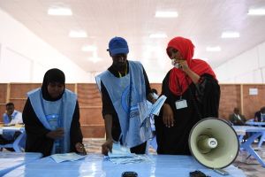 Eine Gruppe von Menschen steht um einen Tisch mit einem Megafon, Papieren und anderen Gegenständen, während andere im Hintergrund auf Stühlen sitzen oder stehen, unter einer Decke mit Lampen, was auf eine Wahllokalität hinweist, in der Menschen ihre Stimmen abgeben.