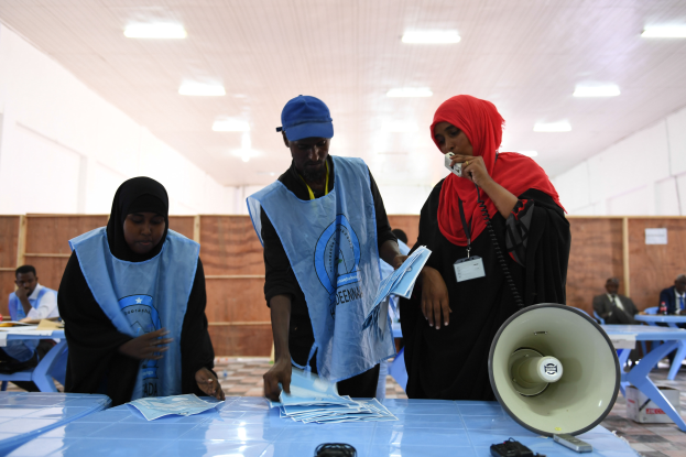 Eine Gruppe von Menschen steht um einen Tisch mit einem Megafon, Papieren und anderen Gegenständen, während andere im Hintergrund auf Stühlen sitzen oder stehen, unter einer Decke mit Lampen, was auf eine Wahllokalität hinweist, in der Menschen ihre Stimmen abgeben.