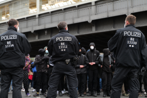 Eine Gruppe von Polizeibeamten in Uniform steht vor einer Menge von Menschen, die ebenfalls schwarze Uniformen und Masken tragen, mit einer Brücke und einem Gebäude im Hintergrund, was auf eine städtische Umgebung hindeutet.