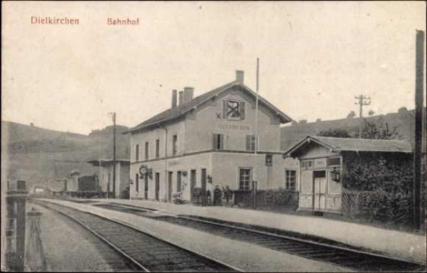 Schwarzes und weißes Foto einer Bahnstation in Dielkirchen, Deutschland, mit einem Zug auf den Gleisen, umgeben von Häusern, Strommasten, Bäumen, Pflanzen, Menschen, fernen Hügeln und einem klaren Himmel.