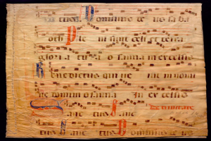 Ein illuminiertes Manuskript mit Musiknotation auf einem Blatt Papier vor einem dunklen Hintergrund, gefüllt mit Text und musikalischen Symbolen, die ein komplexes Design bilden.