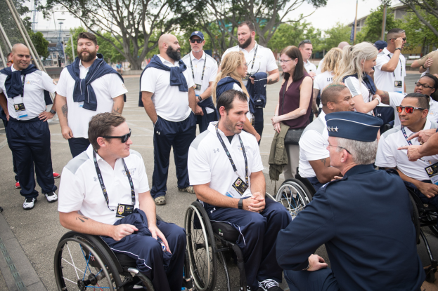 Gruppe von Menschen im Rollstuhl, die sich im Freien unterhalten, vor einem Hintergrund aus Bäumen, Gebäuden und einem klaren blauen Himmel.