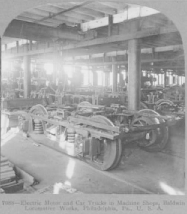 Schwarzes und weißes Foto eines Elektromotors und eines Lkw in einer Fabrik mit Maschinen, Säulen und Dach, mit Text unten.