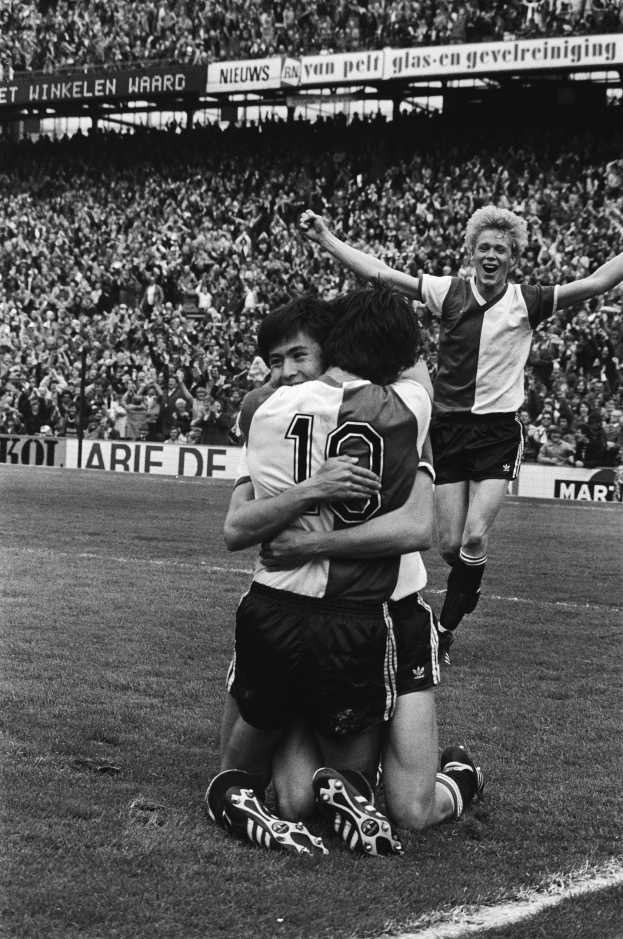 Drei Fußballspieler feiern ein Tor auf dem Boden, mit jubelnder Menge und schwenkenden Fahnen im Hintergrund, auf einem Schwarz-Weiß-Bild.