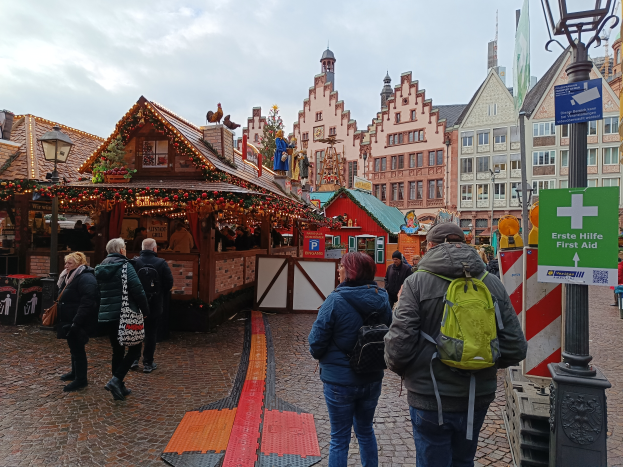 Eine Gruppe von Menschen geht eine Kopfsteinpflasterstraße entlang neben einem Weihnachtsmarkt in Nürnberg, Deutschland, mit Laternenmasten, Texttafeln und Gebäuden mit Fenstern im Hintergrund unter einem bewölkten Himmel.