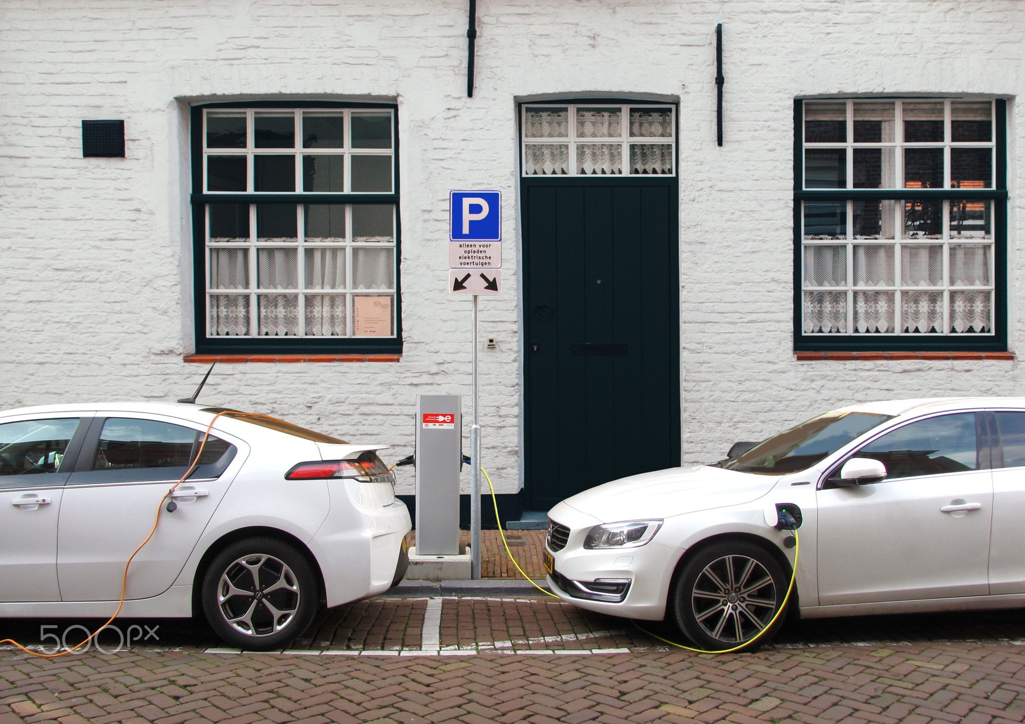 Zwei Elektroautos nebeneinander vor einem weißen Gebäude geparkt, an eine Ladestation angeschlossen.
