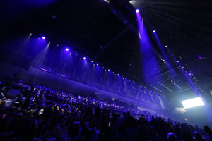 Eine große Menschenmenge, die vor einer Bühne mit Deckenbeleuchtung und einem Bildschirm auf der rechten Seite sitzt, was auf einen Veranstaltungsort hinweist.