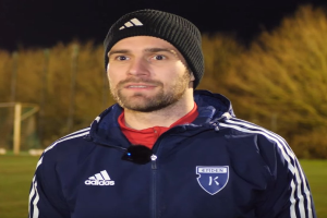 Ein Mann in einer blauen Jacke und Mütze auf einem Fußballfeld während eines Trainings, mit Bäumen und einem Tor im Hintergrund.